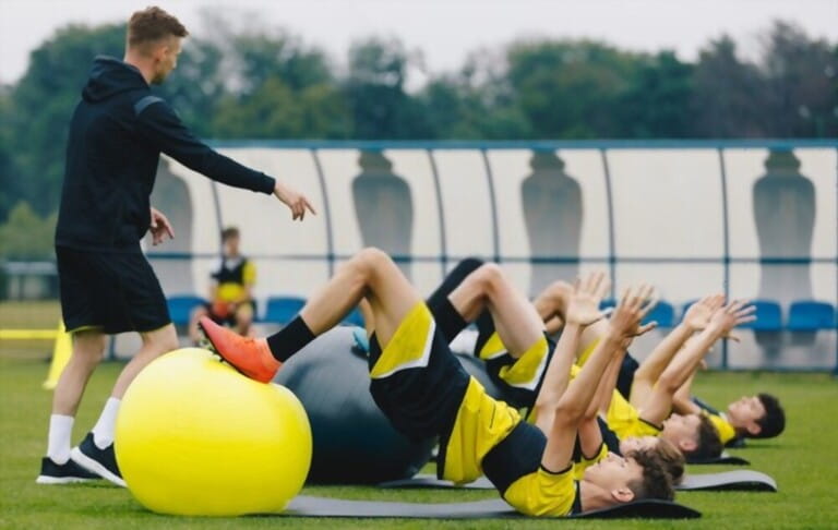 Soccer Players: Stretching Session With Physiotherapist. Youth Sports Team On Recovery Session After League Match. Football Players Warming Up on Grass Stadium Field Soccer Players: Stretching Session With Physiotherapist. Youth Sports Team On Recovery Session After League Match. Football Players Warming Up on Grass Stadium Field