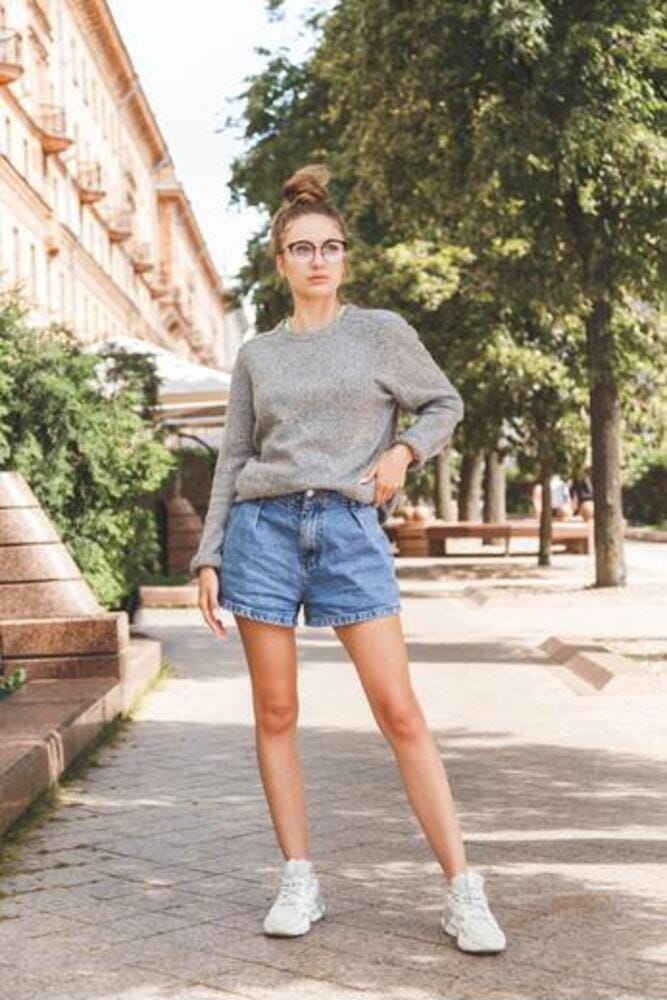Millennial girl in glasses short denim shorts, gray pullover and white sneakers posing on the street. Casual summer clothes of youth. Full length Millennial girl in glasses short denim shorts, gray pullover and white sneakers posing on the street. Casual summer clothes of youth. Full length