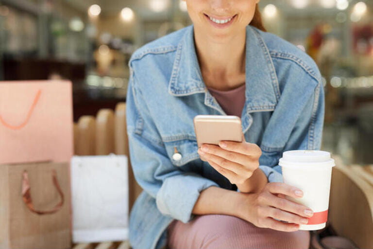 Hands of young smiling girl sitting on bench in mall in purple dress and denim jacket holding coffee Hands of young smiling girl sitting on bench in mall in purple dress and denim jacket holding coffee